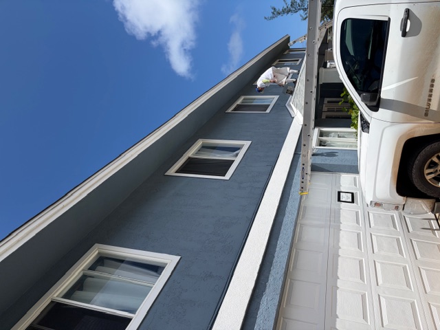 Crew member on a ladder working a two-story residential exterior in blue paint, white pickup parked alongside, mid-coat application